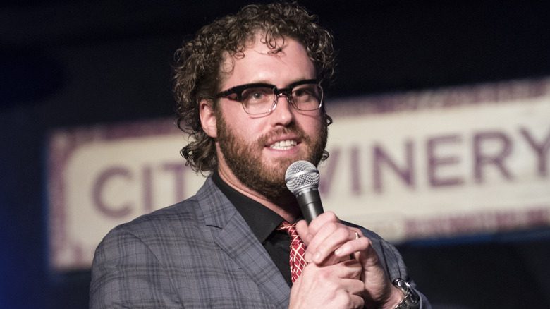 T.J. Miller en costume, portant des lunettes et tenant un microphone