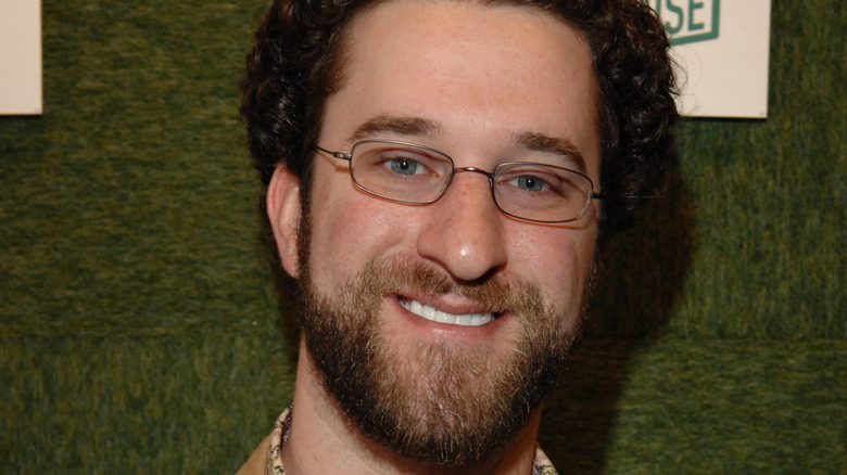 Dustin Diamond avec lunettes et barbe, devant un mur vert