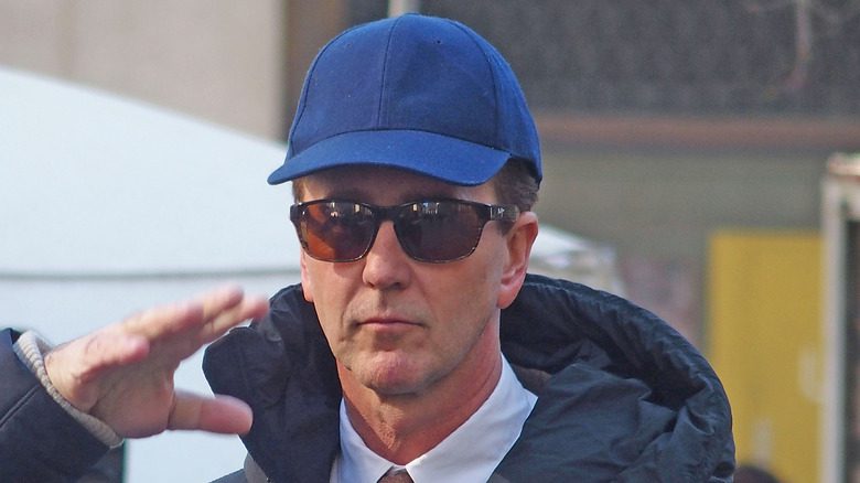 Edward Norton avec une casquette bleue et des lunettes de soleil levant le bras