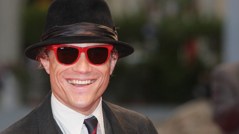 Heath Ledger avec un chapeau trilby, lunettes de soleil rouges, costume, souriant