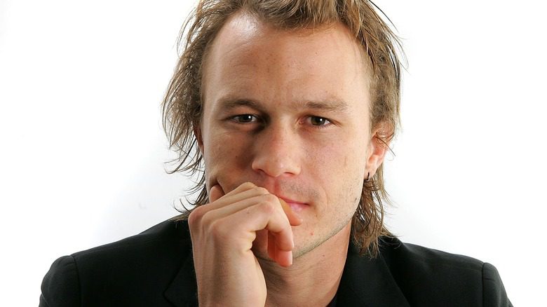 Heath Ledger en costume noir avec la main sur le menton sur fond blanc