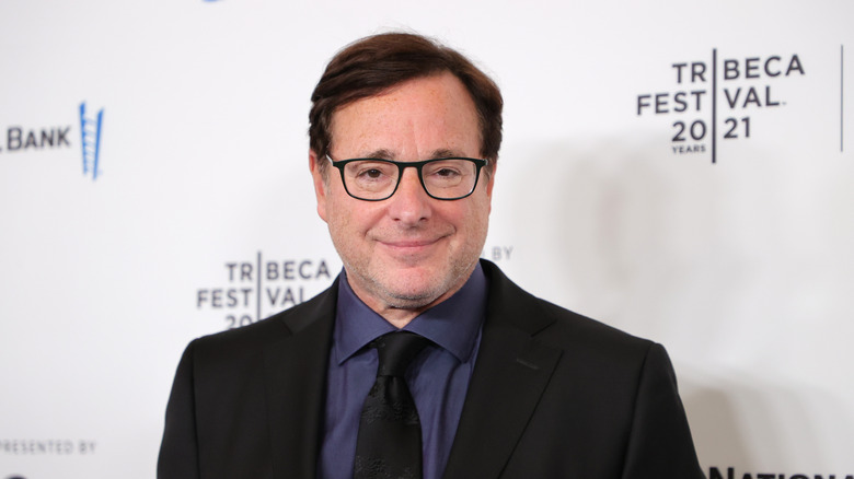 Bob Saget souriant avec des lunettes, costume foncé et cravate lors d'un événement