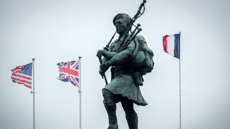 Mémorial de Bill Millin sur Sword Beach en Normandie, avec des drapeaux et la statue de Bill Millin jouant de la cornemuse.