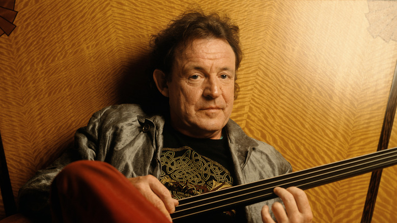 Jack Bruce lying in bed playing guitar smirking at camera